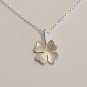 May include: A silver-colored four-leaf clover pendant hangs from a delicate chain. The clover has a textured surface and is centered against a white background. The necklace is a simple, elegant piece of jewelry.