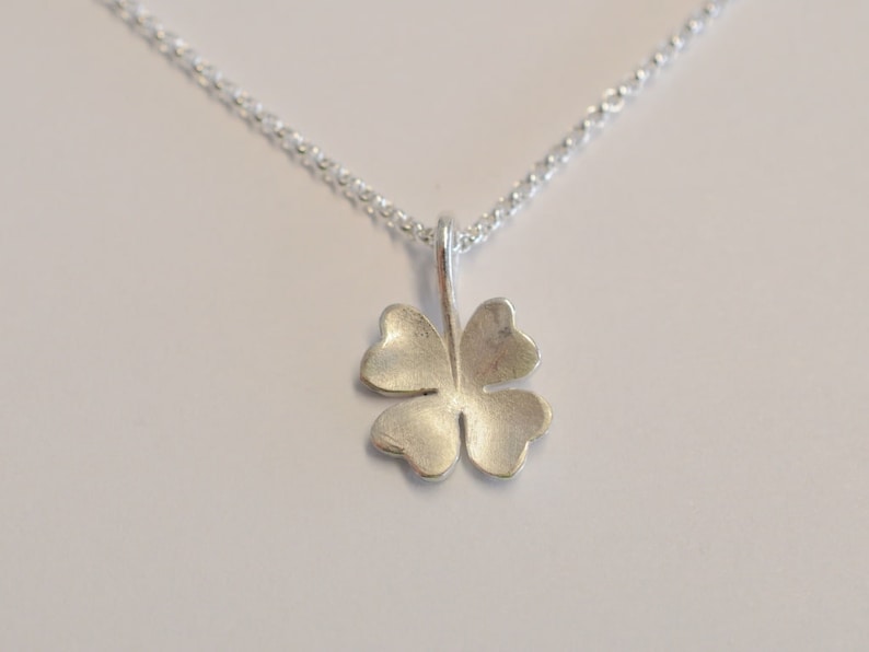 May include: A silver-colored four-leaf clover pendant hangs from a delicate chain. The clover has a textured surface and is centered against a white background. The necklace is a simple, elegant piece of jewelry.