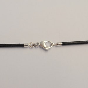 May include: A black leather cord necklace with a silver-colored clasp. The necklace is displayed against a white background. The clasp is a lobster claw style.