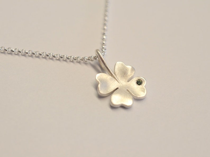 Op de afbeelding: Een zilveren ketting met een klavertje vier hanger met een kleine groene edelsteen in het midden.