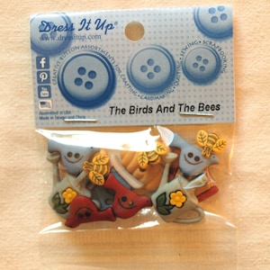 Puede incluir: Un paquete de botones de plástico de colores con pájaros, abejas y regaderas. El paquete está etiquetado como "The Birds And The Bees" y forma parte de la línea de surtidos de botones creativos Dress It Up.