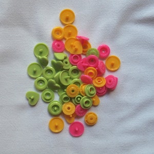 May include: A selection of colourful snap fasteners in yellow, pink, and green. The fasteners are in various shapes, including round and heart-shaped, and are scattered on a white fabric background. These are ideal for sewing and craft projects.