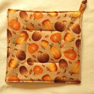 Könnte beinhalten: Ein quadratischer, beigefarbener Topflappen mit einem sich wiederholenden Muster aus orangefarbenen und braunen Kürbissen und Herbstblättern. Der Topflappen hat eine kleine Schlaufe zum Aufhängen. Das Design ist mit einem gesteppten Gittermuster versehen.