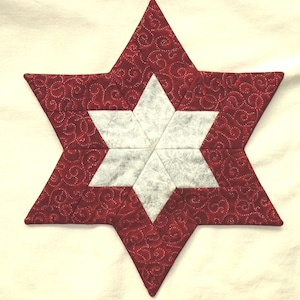 May include: A quilted star-shaped decoration with a red patterned exterior and a white, star-shaped interior. The red fabric features a swirling design. The star is likely handmade and sits on a cream-colored surface.