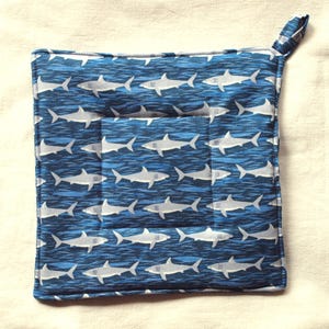 Peut inclure: Un dessous de plat carré bleu et blanc avec un motif de requins. Le dessous de plat est doté d'une boucle pour l'accrocher et est fait de tissu matelassé. Les requins sont blancs et gris, sur un fond bleu.