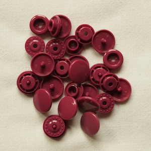 Può includere: Una collezione di bottoni a pressione color bordeaux, in varie dimensioni e stili, sparsi su un tessuto di colore chiaro. I bottoni a pressione includono sia i componenti maschio che femmina, alcuni con dettagli decorativi.