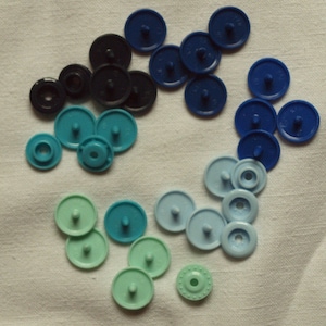 Może przedstawiać: Zestaw plastikowych zatrzasków w różnych odcieniach niebieskiego i zielonego. Zatrzaski są ułożone w półkole na białym tle z tkaniny.
