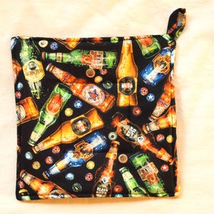 May include: A black square pot holder with a loop for hanging. The pot holder is covered in a pattern of beer bottles and bottle caps in various colors, including green, gold, and blue. The bottles are arranged in a random pattern.