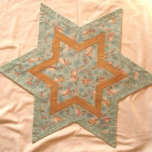 Puede incluir: Un mantel individual acolchado en forma de estrella. El centro de la estrella es de color marrón cálido, con un fondo azul claro con un estampado floral de pájaros. Los bordes son de un verde azulado más oscuro. Este mantel individual decorativo añade encanto.