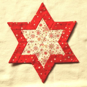May include: A red and white quilted star-shaped decoration. The star has a red border with gold stars and a center with a white background and a pattern of red and gold snowflakes. A festive holiday decoration.
