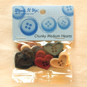 Op de afbeelding: Een verpakking met dikke, middelgrote hartvormige knopen in verschillende kleuren, waaronder bruin, rood, beige en groen. De verpakking is gelabeld met "Dress It Up" en "Chunky Medium Hearts".