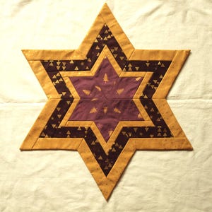 Könnte beinhalten: Ein handgefertigter sternförmiger Quilt mit geometrischem Design. Der Stern hat eine tiefviolette Mitte mit goldenen Baummustern, umgeben von dunkelbraunen und goldenen Dreiecken und einem goldgelben Rand. Das Objekt ist wahrscheinlich ein dekorativer Artikel.