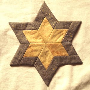 Op de afbeelding: Een handgemaakt, stervormig quiltblok met zes punten. De ster heeft een goudgeel midden en een getextureerde, grijze rand. De grijze stof heeft een subtiel patroon met gouden spikkels.