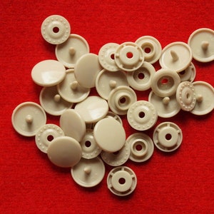 May include: A collection of beige plastic snap buttons, some with a raised center post, others with a flat top.