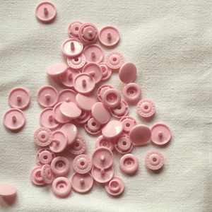 Peut inclure: Un tas de boutons-pression en plastique rose sur un fond de tissu blanc.