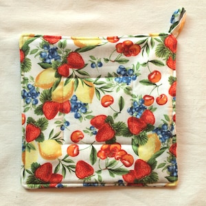 Könnte beinhalten: Ein weißer Baumwoll-Topflappen mit einem bunten Fruchtmuster. Das Muster enthält Erdbeeren, Zitronen, Kirschen und Blaubeeren. Der Topflappen hat einen Schlaufe.