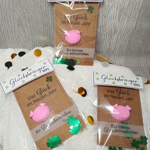 May include: Three small packages of soap, each with a pink pig-shaped soap and a tag that reads "Glücksbringer SEIFE" and "Viel Glück im Neuen Jahr". The packages are on a white, fluffy surface with gold confetti.