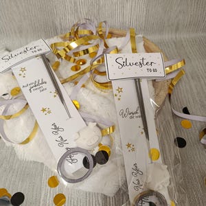 Puede incluir: Dos bolsas de regalo con el texto "Silvester TO 60" y "New Year" en tarjetas blancas. Cada bolsa contiene un petardo, una pequeña figura de cerdo y un anillo. Confeti y cintas doradas y plateadas rodean las bolsas.