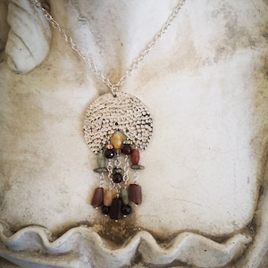 Könnte beinhalten: Eine silberne Halskette mit einem runden Anhänger mit gehämmerter Textur und einem herabhängenden Cluster aus mehrfarbigen Edelsteinen, darunter Braun, Grün, Gelb und Rot.