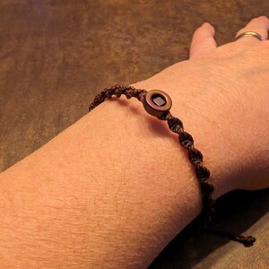 May include: A brown braided bracelet with a round, dark brown bead with a square centre.