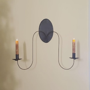 May include: A black metal wall sconce with two candle holders. The sconce features an oval backplate and curved arms that hold the candle holders. The candles are brown with a flickering yellow light.