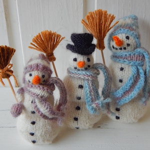 Könnte beinhalten: Drei handgestrickte weiße Schneemannfiguren, jede mit einer leuchtend orangefarbenen Karottennase, schwarzen gestickten Augen und Knöpfen, einem bunten Garnschal und einem kleinen Holzbesen mit orangefarbenen Borsten.