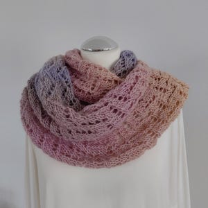 Könnte beinhalten: Ein handgestrickter Cowl-Schal in einem Farbverlauf aus Rosa, Lavendel und Pfirsich. Das Lochmuster des Schals ist sichtbar, und er wird auf einem weißen Mannequinkopf präsentiert. Der Schal ist aus weichem Garn gefertigt.