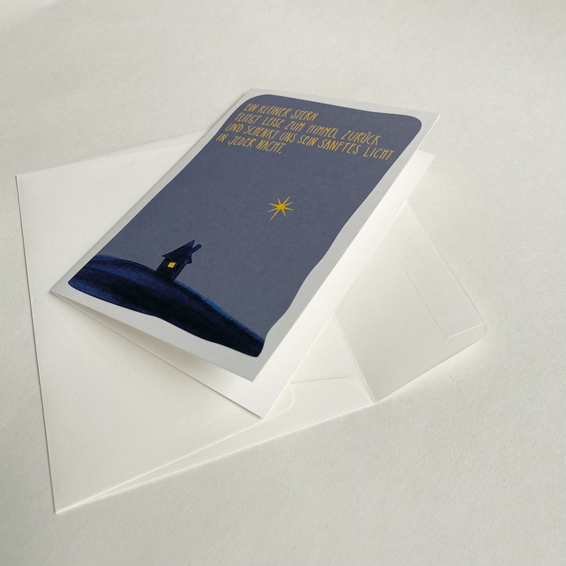 May include: A greeting card with a blue night sky background, a small house with a lit window, and a yellow star. The text on the card reads "Ein kleiner Stern fliegt leise zum Himmel zur&uuml;ck und schenkt uns sein sanftes Licht in jeder Nacht."