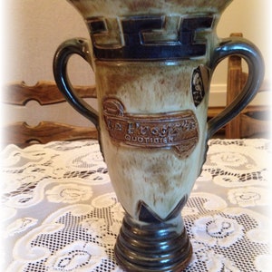 Peut inclure: Un vase en céramique brun et vert avec deux anses. Le vase a un motif décoratif autour du haut et une étiquette qui indique "La Propose Quotidien".