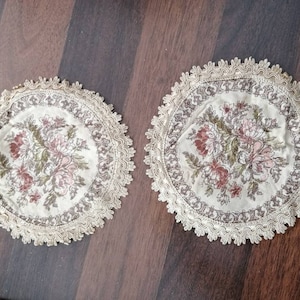 Könnte beinhalten: Zwei runde, weiße, gehäkelte Deckchen mit einem Blumenmuster in Rosa-, Braun- und Grüntönen. Die Deckchen haben einen gewellten Rand.