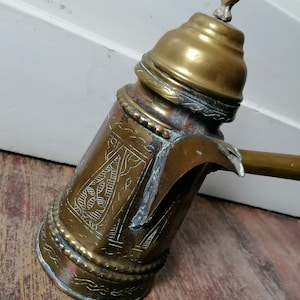 Puede incluir: Una cafetera de latón vintage con intrincados diseños grabados. La cafetera tiene un cuerpo redondeado y un pico con asa. La parte superior de la cafetera tiene un remate decorativo.