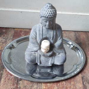 Könnte beinhalten: Eine graue Steinbuddha-Statue, die in meditativer Haltung sitzt und eine weiße Kerze auf ihrem Schoß hält. Die Statue steht auf einem silbernen Tablett mit einem dekorativen Rand.