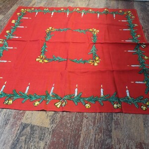 May include: A red Christmas tablecloth with a festive design. The border features green garland, white candles, and gold bells. A smaller square in the centre repeats the garland and bell motif. The tablecloth is made of a woven fabric.