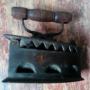 Puede incluir: Una plancha de hierro fundido vintage con mango de madera. La plancha tiene una serie de agujeros en la parte inferior y un diseño decorativo en el lateral.