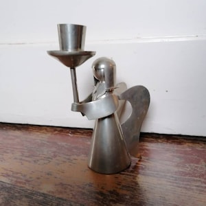 May include: A silver metal angel candle holder with wings and a candle cup on top. The angel is holding the candle cup in its right hand.