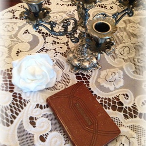 May include: A silver candelabra with four candle holders sits on a white lace tablecloth. A white rose and a brown leather-bound journal are also on the table.
