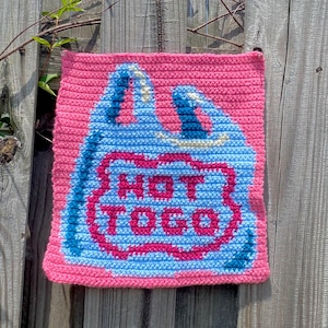 May include: A pink crocheted square bag with a blue and white design resembling a shopping bag. The bag has the words "HOT TO GO" in a red outline. The bag is hanging on a weathered wooden fence with green vines.