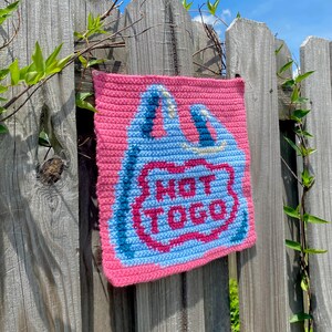 K&ouml;nnte beinhalten: Ein rosa geh&auml;keltes Quadrat mit einem blau-rosa Design, das einer Einkaufstasche &auml;hnelt. Der Text "NOT TOGO" ist in der Mitte der Tasche zu sehen. Das Quadrat h&auml;ngt an einem verwitterten Holzzaun mit gr&uuml;nen Ranken.