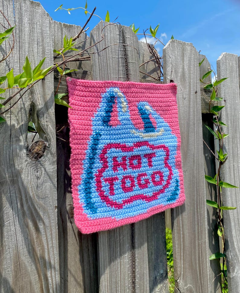 K&ouml;nnte beinhalten: Ein rosa geh&auml;keltes Quadrat mit einem blau-rosa Design, das einer Einkaufstasche &auml;hnelt. Der Text "NOT TOGO" ist in der Mitte der Tasche zu sehen. Das Quadrat h&auml;ngt an einem verwitterten Holzzaun mit gr&uuml;nen Ranken.