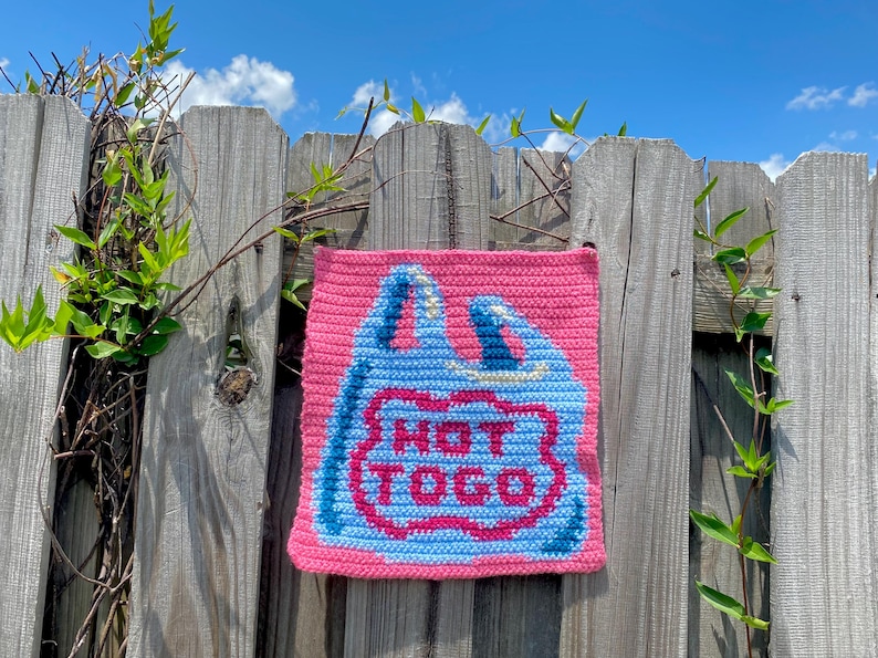 K&ouml;nnte beinhalten: Eine pinkfarbene, geh&auml;kelte quadratische Tasche mit einem blau-wei&szlig;en Design, das einer Einkaufstasche &auml;hnelt. Die Tasche hat die Worte "HOT TO GO" in roter Umrandung. Die Tasche h&auml;ngt an einem verwitterten Holzzaun mit gr&uuml;nen Ranken.