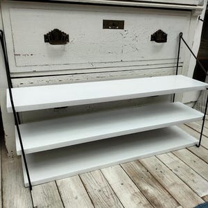 May include: A white, three-tiered floating shelf with black metal supports. The shelves are rectangular and mounted against a distressed white cabinet. The shelves are empty and ready for display.