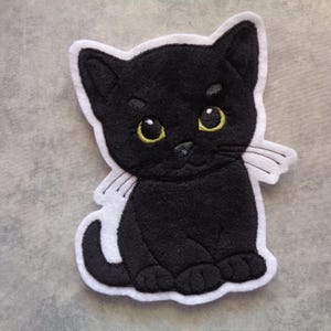 Könnte beinhalten: Ein gestickter Aufnäher in Form einer sitzenden schwarzen Katze mit großen gelben Augen und weißen Schnurrhaaren. Die Katze hat einen einfarbig schwarzen Körper mit einem dicken weißen Rand, der die Silhouette betont und detaillierte Stickereien aufweist.