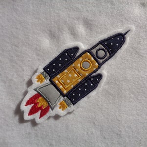 Könnte beinhalten: Eine blaue und gelbe Rakete mit weißen Sternen und einer roten Flamme, gestickt auf einem weißen Stoff.