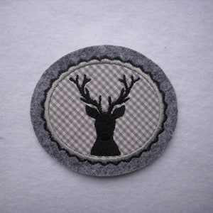 Könnte beinhalten: Ein grauer Filzuntersetzer mit einer schwarzen gestickten Hirschsilhouette auf einem beige-weißen Gingham-Hintergrund.
