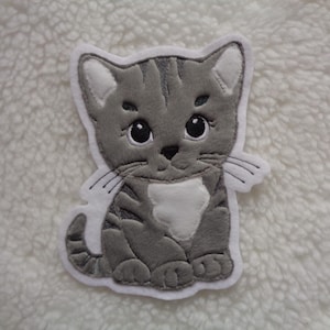 Könnte beinhalten: Ein gestickter Aufnäher in Form einer grau-weißen Katze, der ein Kätzchen im Cartoon-Stil mit großen schwarzen Augen, dunkelgrauen Streifen, einem weißen Brustfleck und weißen Innenohren zeigt, eingefasst mit einem weißen Filzrand.