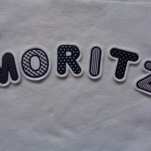 Könnte beinhalten: Ein Filznamensschild mit dem Namen "MORITZ" in schwarzen und weißen gemusterten Buchstaben.