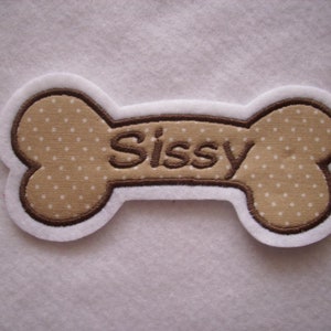 Könnte beinhalten: Ein brauner und weißer, knochenförmiger Aufnäher mit dem Namen "Sissy", der in braunem Garn gestickt ist.