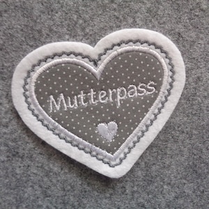 Könnte beinhalten: Ein weißer Filz-Herz-Patch mit einem grauen Herz mit weißen Punkten darin. Das graue Herz hat weiße Stickerei mit der Aufschrift "Mutterpass".