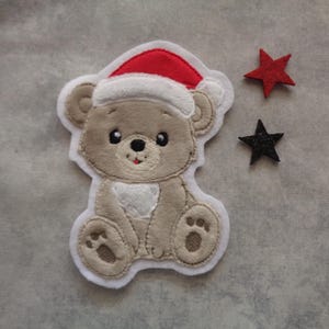 Könnte beinhalten: Ein gestickter Aufnäher mit einem Cartoon-Teddybär, der einen Weihnachtsmannhut trägt. Der Bär ist hellbraun mit weißem Bauch und Pfoten. Zwei sternförmige Verzierungen, eine rote und eine schwarze, sind ebenfalls vorhanden.
