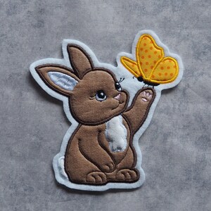 Könnte beinhalten: Ein braunes Kaninchen mit weißem Fell auf der Brust schaut ein gelbes Schmetterling mit orangefarbenen Punkten an.